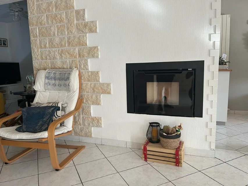 Intérieur chaleureux avec cheminée électrique blanche allumée, fauteuil gris moderne, étagères en bois avec bûches décoratives